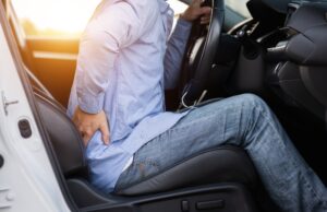 A man in the driver's seat of a car, showing signs of discomfort from back pain.