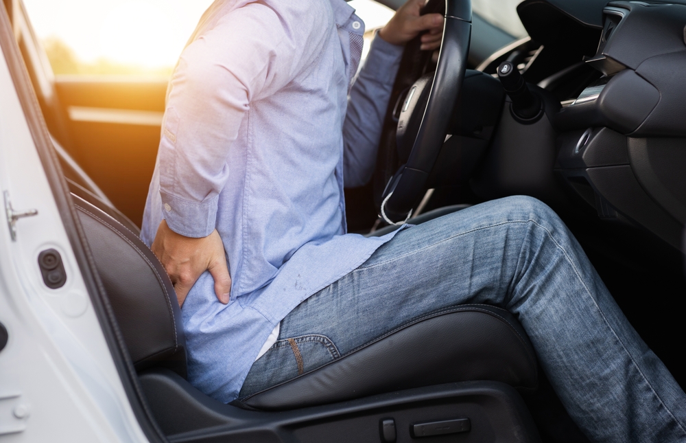 A man in the driver's seat of a car, showing signs of discomfort from back pain.